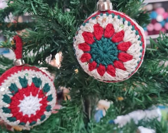 May include: Two handmade Christmas ornaments with a crocheted design. The ornaments are round, with a red, white, and green floral pattern. They hang from a green artificial Christmas tree. The ornaments have a silver metal cap and a loop for hanging.