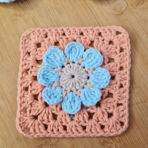 May include: A crocheted square with a light blue flower in the center. The square is made of peach-colored yarn and the flower is made of light blue yarn.