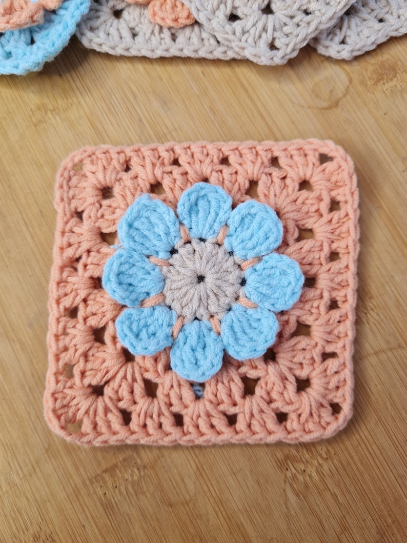 May include: A crocheted square with a light blue flower in the center. The square is made of peach-colored yarn and the flower is made of light blue yarn.