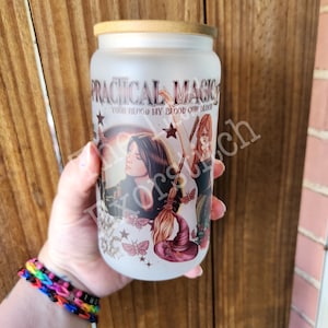 May include: Frosted glass tumbler with a bamboo lid. The tumbler features the text "PRACTICAL MAGIC" and an illustration of witches, a broom, and a hat. The design includes stars and butterflies. The tumbler is held by a person.