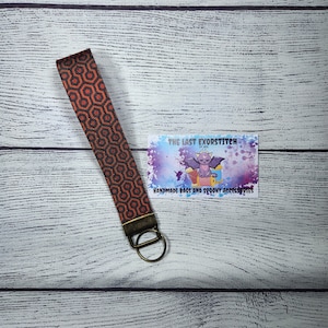 May include: A black and orange patterned key chain with a brass key ring. The keychain is on a white wooden surface. The business card reads "The Last Exorstitch Handmade Bags and Spooky Accessories."