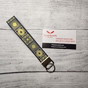 May include: A black and yellow key chain with a geometric pattern. The key chain is attached to a silver key ring. The key chain is on a white wooden surface. The Last Exorstitch - EST. 2022 - HANDMADE WALLETS AND BAGS WITH A SPOOKY TWIST! Custom requests welcomed Thelastexorstitch.com Facebook.com/thelestexorstitch