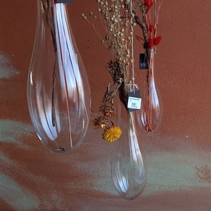 Könnte beinhalten: Drei klare Glasvasen mit getrockneten Blumen, die an einer Holzstange hängen. Die Vasen sind tropfenförmig und haben eine strukturierte Oberfläche. Die Blumen sind eine Mischung aus getrockneten Gräsern, roten Blumen und kleinen gelben Blumen.