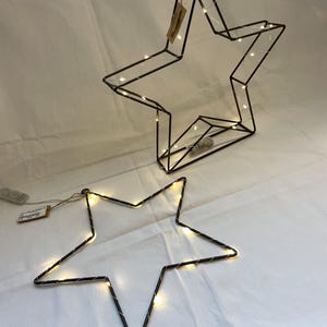 Könnte beinhalten: Zwei sternförmige Leuchten aus schwarzem Metall mit warmweißen LED-Lichtern. Der größere Stern steht aufrecht, während der kleinere Stern flach liegt.