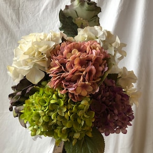 Könnte beinhalten: Ein Strauß künstlicher Hortensien in den Farben Weiß, Grün, Rosa und Lila. Die Blumen sind in einem handgehaltenen Strauß angeordnet.