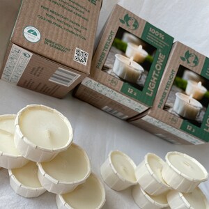 Könnte beinhalten: Eine Schachtel mit 12 weißen, unparfümierten Papier-Teelichtern. Die Schachtel ist aus Pappe und hat ein grün-weißes Logo mit dem Text "LIGHT.ONE" und "100% NATURWACHS". Die Teelichter bestehen aus natürlichem Wachs und sind feuerfest. Die Schachtel wird in Deutschland hergestellt.