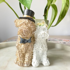 May include: A figurine of a brown dog wearing a top hat and bow tie standing next to a white dog wearing a flower crown. The dogs are on a white surface.