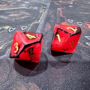 May include: Two red and black dice with gold numbering. The dice are six-sided and have the numbers 1, 2, 3, and 4 visible. The dice are on a textured surface.