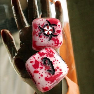May include: Two dice with a white and red splattered design. One die features a black and white biohazard symbol, the other shows a black number 2. The dice are held in a hand.