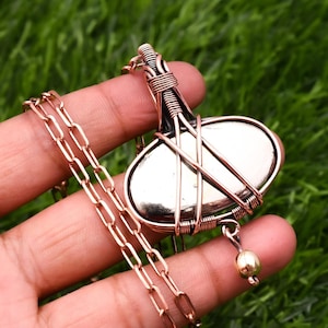 May include: A silver oval-shaped stone pendant wrapped in copper wire with a copper chain. The pendant has a small gold bead hanging from the bottom.