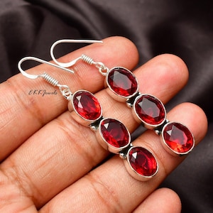 May include: A pair of dangle earrings featuring three oval-cut, deep red garnet gemstones set in polished silver metal. Each earring has a hook-style closure and displays the text EKKJewels on the side. The stones are faceted and arranged vertically.