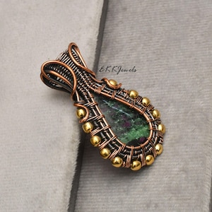 May include: A teardrop-shaped pendant featuring a dark green and magenta stone encased in intricate copper wire wrapping. The design includes small gold-toned beads along the outer edge and the text EKKJewels printed in black near the top.