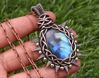 Puede incluir: Un colgante envuelto en alambre de cobre con una piedra preciosa de labradorita azul. El colgante está rodeado por un diseño de alambre de cobre con detalles dorados. El colgante está unido a una cadena de cobre.