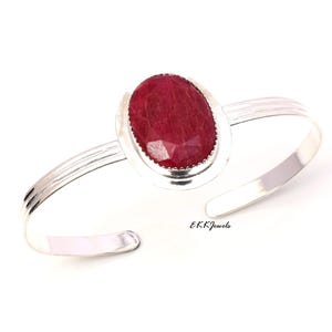 Può includere: Un bracciale rigido in argento con una grande pietra preziosa rosso scuro, di forma ovale e sfaccettata, incastonata in una lunetta di metallo liscio. Il bracciale ha un design a tre linee ed è aperto. Il testo EKKJewels è stampato in nero.