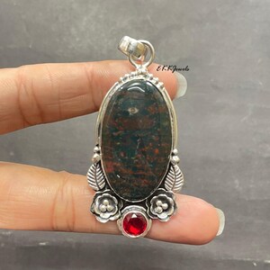 May include: An oval-shaped pendant featuring a dark green and reddish-brown mottled stone set in a silver frame. The setting includes decorative silver leaves and flowers, with a small, round, faceted red gemstone at the base and a silver bail.