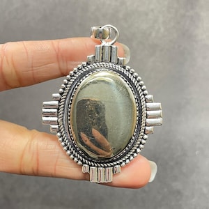 Könnte beinhalten: Silberner Anhänger mit einem ovalen Pyrit-Stein, der in einen dekorativen Rahmen gefasst ist. Der Rahmen hat ein geometrisches Design mit einem seilartigen Rand.