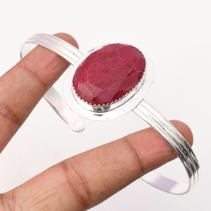 Könnte beinhalten: Ein silbernes Armreif-Armband mit einem roten ovalen Rubin-Edelstein, der in einer silbernen Fassung sitzt. Das Armband ist offen und verstellbar.