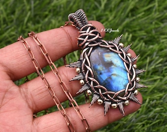 Può includere: Un ciondolo in filo di rame avvolto con una pietra di labradorite ovale dai riflessi blu, incastonata in filo di rame con piccole perline metalliche e punte radianti, fissato a una catena di rame. Il testo EKKJewels è visibile sul ciondolo.
