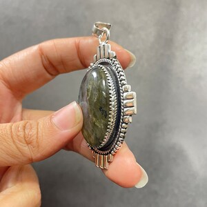 Puede incluir: Un colgante de plata con una gran piedra de labradorita de forma ovalada. El colgante tiene un dise&ntilde;o de plata detallado alrededor de la piedra y una anilla en la parte superior para sujetar una cadena.