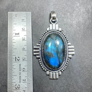 Puede incluir: Un colgante de plata con una gran piedra ovalada de labradorita engastada en un marco de plata. El marco tiene un dise&ntilde;o geom&eacute;trico con forma de cruz. El colgante mide aproximadamente 5 cm de largo.