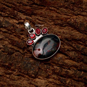 Peut inclure: Un pendentif en argent orn&eacute; d'une grande pierre ovale sombre aux inclusions rouge&acirc;tres, rehauss&eacute; de quatre petites pierres rouges ovales dispos&eacute;es en &eacute;ventail au-dessus. La monture m&eacute;tallique comporte une b&eacute;li&egrave;re avec le texte E K K Jewels.