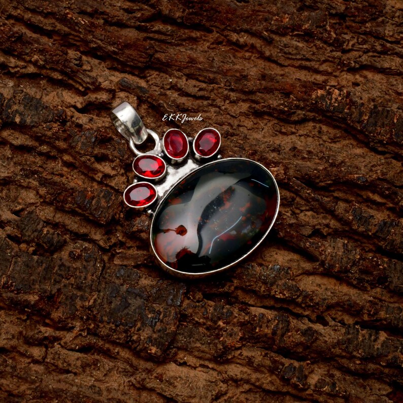 Peut inclure: Un pendentif en argent orn&eacute; d'une grande pierre ovale sombre aux inclusions rouge&acirc;tres, rehauss&eacute; de quatre petites pierres rouges ovales dispos&eacute;es en &eacute;ventail au-dessus. La monture m&eacute;tallique comporte une b&eacute;li&egrave;re avec le texte E K K Jewels.