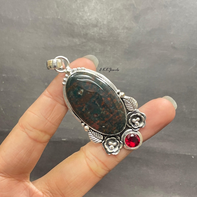 May include: A silver pendant featuring a large oval bloodstone with dark green and red mottled patterns. The setting includes decorative silver leaves, two small silver flowers with beaded centers, and a single round faceted red gemstone.