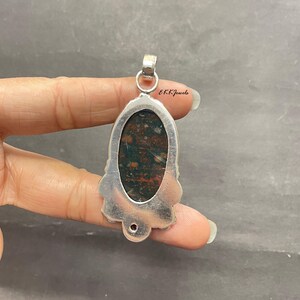 May include: A silver pendant featuring an oval-shaped bloodstone with dark green and red speckled patterns. The metal setting has a decorative scalloped edge and a circular bail at the top. The text EKKJewels is printed near the top of the pendant.