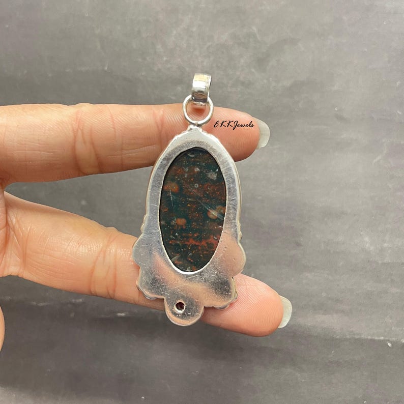May include: A silver pendant featuring an oval-shaped bloodstone with dark green and red speckled patterns. The metal setting has a decorative scalloped edge and a circular bail at the top. The text EKKJewels is printed near the top of the pendant.