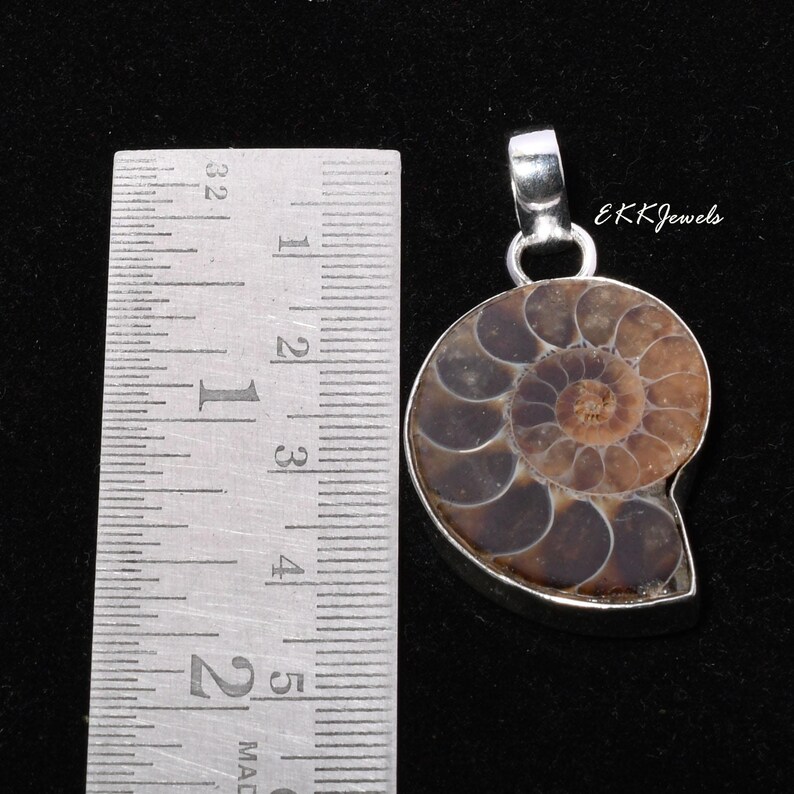 Puede incluir: Un colgante de f&oacute;sil de amonita en espiral marr&oacute;n y bronceado engastado en un marco de metal plateado con una anilla, colocado junto a una regla de metal que muestra una altura de unos 3,8 cm para la pieza f&oacute;sil, etiquetado EKKJewels.