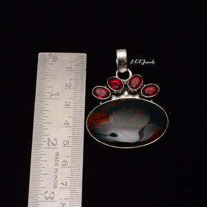 Peut inclure: Un pendentif en argent avec une grande pierre d'ambre ovale fonc&eacute;e et quatre petites pierres grenat rouges ovales, plac&eacute; &agrave; c&ocirc;t&eacute; d'une r&egrave;gle m&eacute;tallique indiquant une longueur d'environ 3,8 cm. Le texte EKKJewels est visible sur le pendentif.