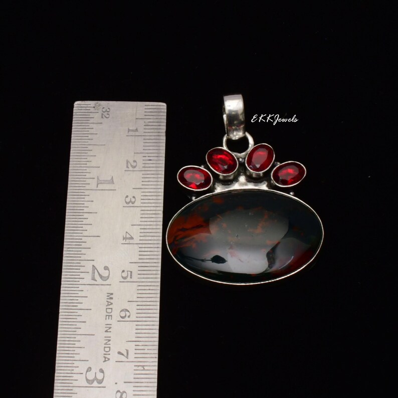 Peut inclure: Un pendentif en argent avec une grande pierre d'ambre ovale fonc&eacute;e et quatre petites pierres grenat rouges ovales, plac&eacute; &agrave; c&ocirc;t&eacute; d'une r&egrave;gle m&eacute;tallique indiquant une longueur d'environ 3,8 cm. Le texte EKKJewels est visible sur le pendentif.