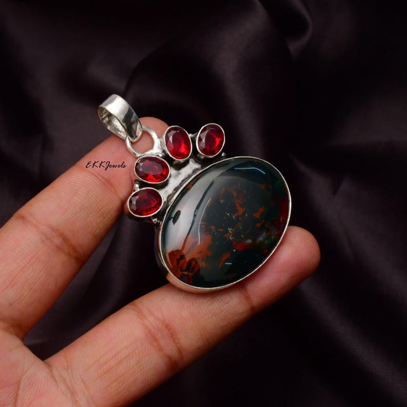 Peut inclure: Un pendentif en argent avec une grande pierre ovale vert fonc&eacute; et brun rouge&acirc;tre sertie dans une monture en m&eacute;tal poli. Le haut du pendentif est orn&eacute; de quatre petites pierres pr&eacute;cieuses rouges taille ovale. Le texte EKKJewels est inscrit sur le m&eacute;tal.