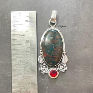 May include: An oval bloodstone pendant set in silver with floral and leaf accents, featuring a small round red gemstone at the base. A metal ruler placed alongside shows the pendant is approximately 2.5 inches long. Text EKKJewels is visible.