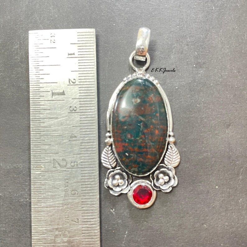 May include: An oval bloodstone pendant set in silver with floral and leaf accents, featuring a small round red gemstone at the base. A metal ruler placed alongside shows the pendant is approximately 2.5 inches long. Text EKKJewels is visible.