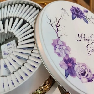 May include: A white round tin box with a purple floral design and the text "Has Geldin Gökcen". The box is filled with small white chocolate squares.