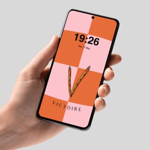 Può includere: Uno smartphone mostra uno sfondo digitale a scacchi nei toni del rosa e dell'arancione. L'ora 19:26 e la data sono visualizzate. La parola "VICTOIRE" è in basso e una forma a "V" fatta di grissini è al centro.