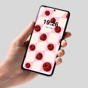 Pode incluir: Um smartphone exibe um padrão xadrez rosa e branco com framboesas vermelho escuro. A hora 19:26 e a data Sáb, 17 de maio são visíveis. O telefone é segurado em uma mão.