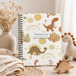 May include: A cream-coloured baby memory book with a spiral binding, decorated with a dinosaur and sun pattern. The text "Ton histoire à toi" is displayed on a white label. A stuffed dinosaur and wooden toy are also visible.