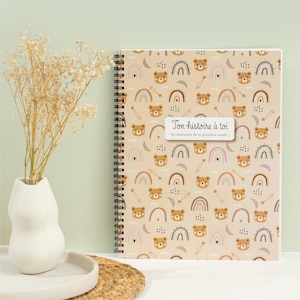 May include: A spiral-bound journal with a beige cover featuring a repeating pattern of brown teddy bear faces, rainbows, and stars. The journal has a white label with the text "Ton histoire à toi". A vase with dried flowers is to the left.