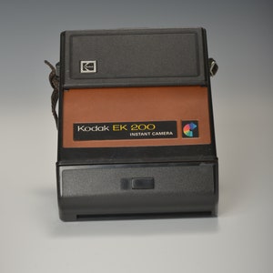 Puede incluir: Una cámara instantánea Kodak EK 200 negra y marrón con una correa. La cámara tiene una parte superior negra y una parte delantera marrón con el texto "Kodak EK 200 Instant Camera" en letras amarillas.