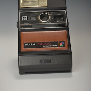 Puede incluir: Una cámara instantánea Kodak EK300 negra y marrón con flash incorporado. La cámara tiene una cubierta de cuero marrón en la parte delantera con las palabras "Kodak EK300 Instant Camera" impresas en blanco.