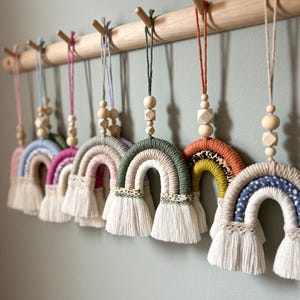 May include: A row of seven colourful rainbow wall hangings with wooden beads and fringe. The rainbows are made of yarn and have different patterns and colours.