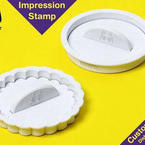 May include: White plastic cookie cutter with a cat design. The cutter has a scalloped edge and a removable handle. The text "Cookie Cat" and "Impression Stamp" are visible in the image.