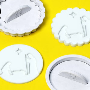 May include: Three white plastic cookie cutters shaped like a nativity scene. The cutters have a star, a manger, and three figures. The cutters have a scalloped edge and a handle on the back. The handle has a black cat face with the text "cookie cat" underneath.