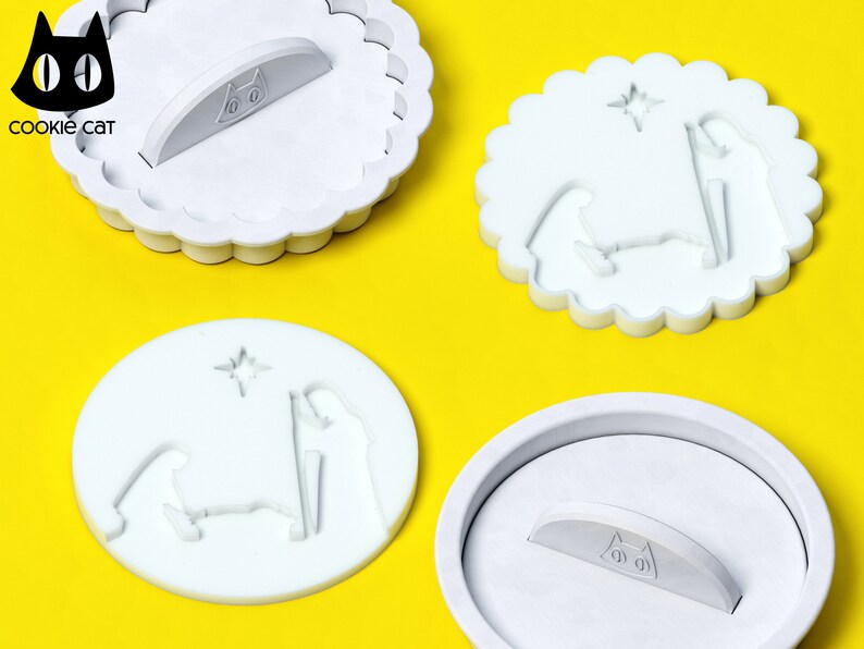 May include: Three white plastic cookie cutters shaped like a nativity scene. The cutters have a star, a manger, and three figures. The cutters have a scalloped edge and a handle on the back. The handle has a black cat face with the text "cookie cat" underneath.