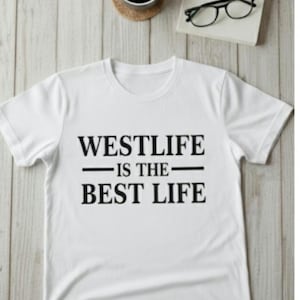 May include: White t-shirt with the text "WESTLIFE IS THE BEST LIFE" in black. The shirt is on a light wood-grain surface, with a cup of coffee and a pair of glasses in the background.