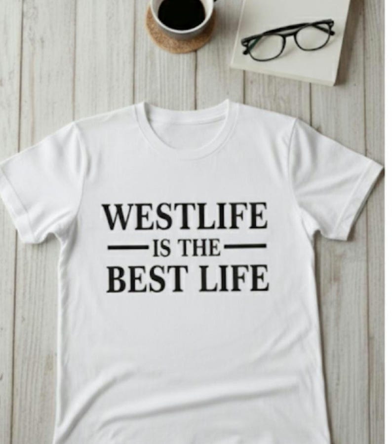 May include: White t-shirt with the text "WESTLIFE IS THE BEST LIFE" in black. The shirt is on a light wood-grain surface, with a cup of coffee and a pair of glasses in the background.