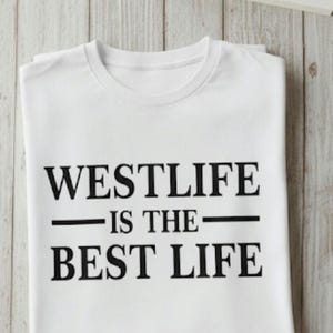 May include: White t-shirt with the text "WESTLIFE IS THE BEST LIFE" in black. The shirt is folded and placed on a light wood-grain surface, with a cup of coffee, a coaster, glasses, and a book in the background.