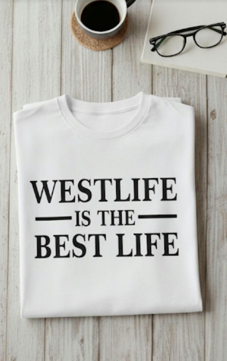 May include: White t-shirt with the text "WESTLIFE IS THE BEST LIFE" in black. The shirt is folded and placed on a light wood-grain surface, with a cup of coffee, a coaster, glasses, and a book in the background.